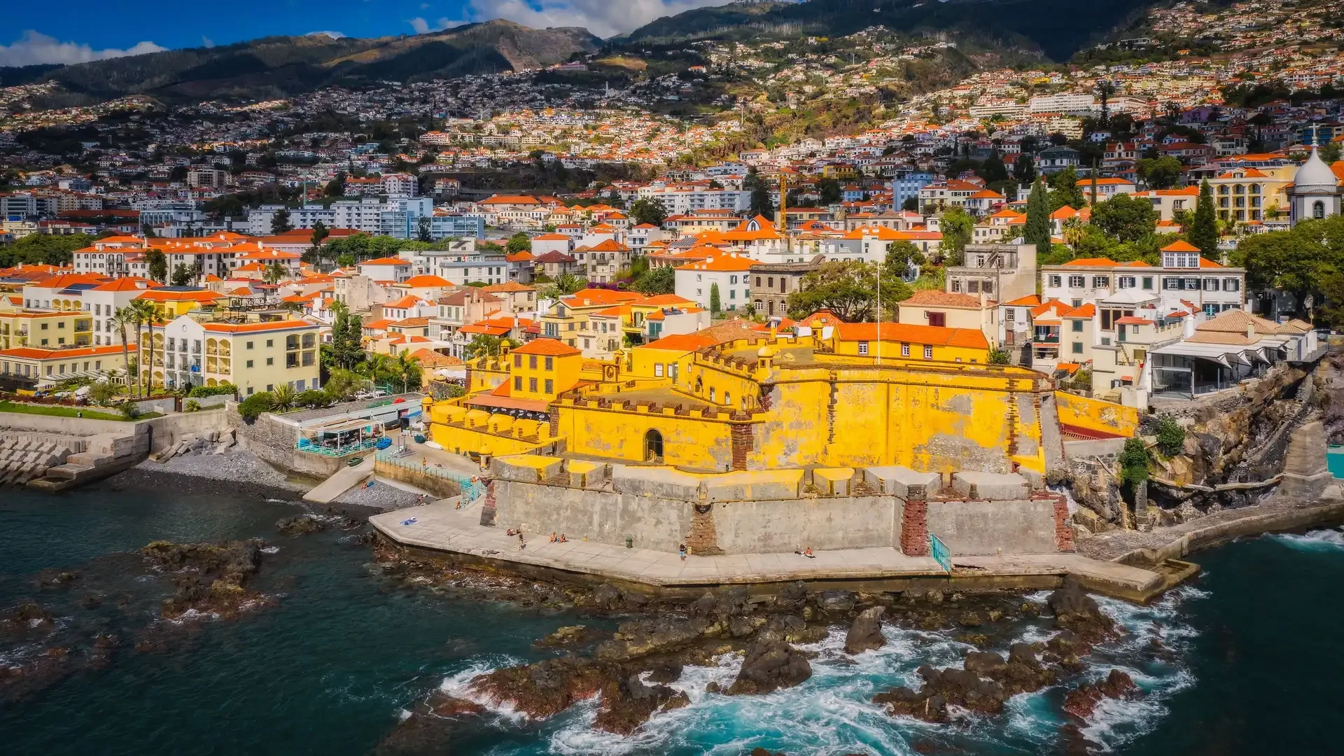 Aerial view of a coastal city with colorful buildings, including a prominent yellow fortress-like structure at the water's edge, and hilly terrain in the background.