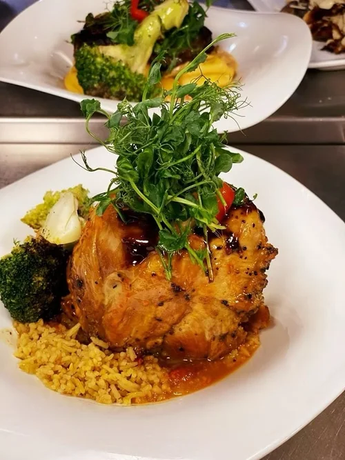 A plate with a roasted chicken thigh topped with fresh greens and sauce, served with broccoli, cauliflower, and yellow rice