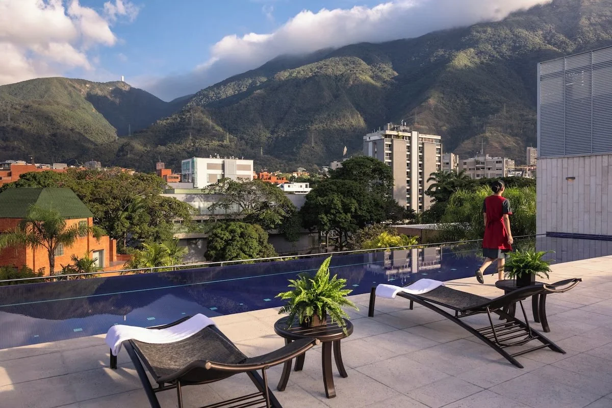 A rooftop view of a city with lush green hills in the background, featuring a swimming pool, lounge chairs with towels, potted plants, and a woman in a red dress walking.