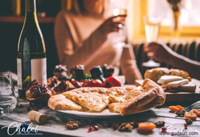 A table with a pizza, cupcakes, strawberries, wine, and cheese with people in the background holding glasses, in a cozy restaurant setting.