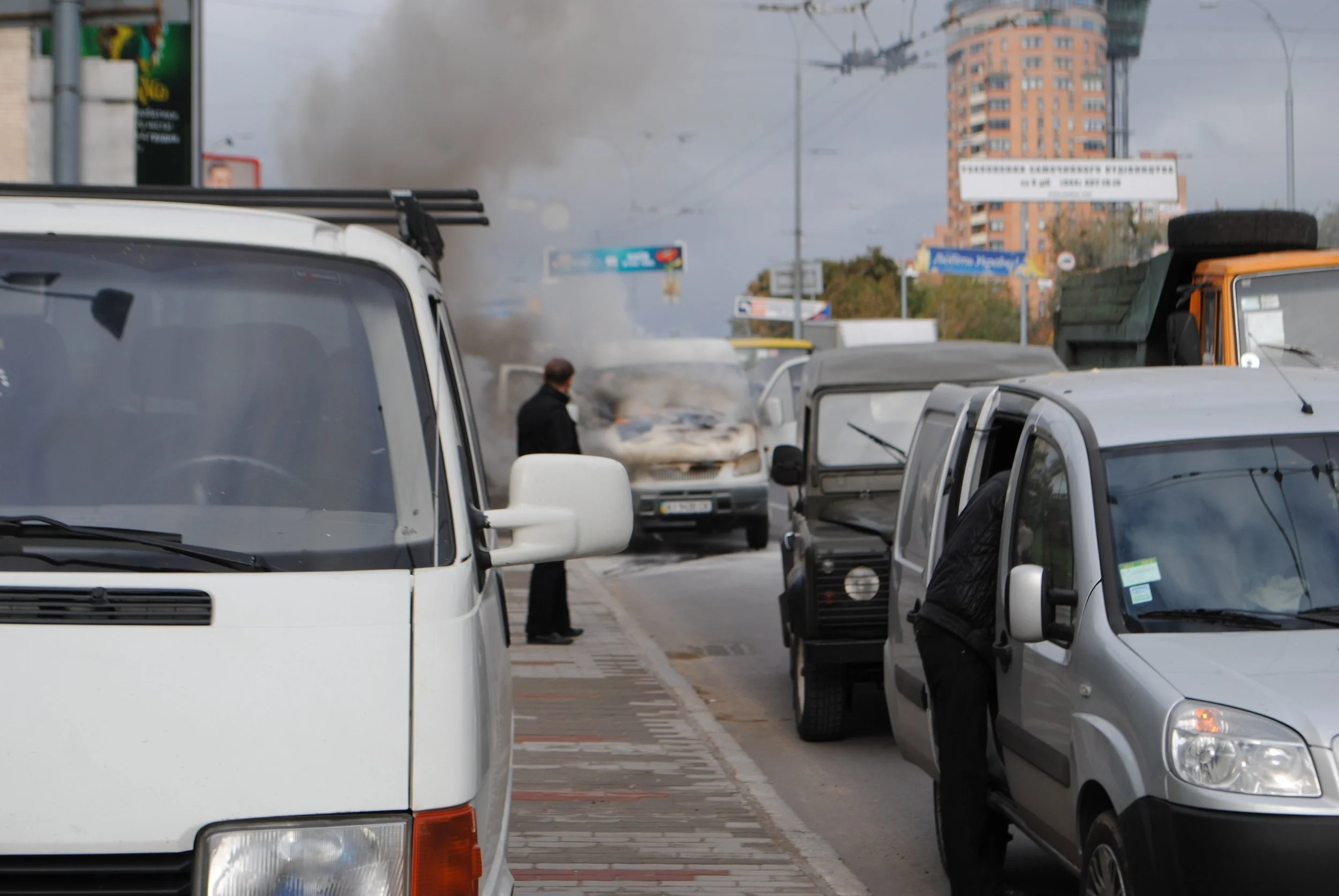 Multiple parked vehicles on the street, smoke or fog in the background with a tall building, and a person standing near the vehicles. Kiev November 2011, Marina Parrish ©