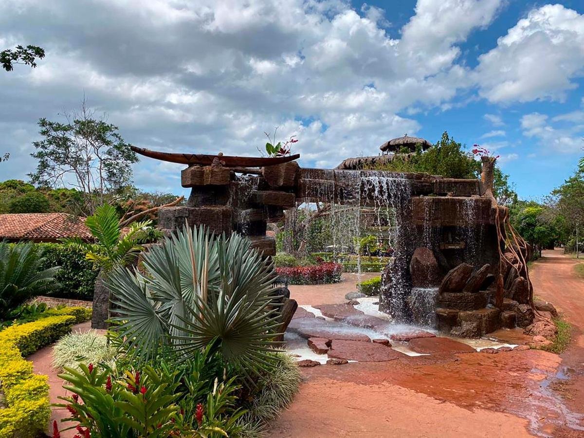 A decorative stone structure with water cascading down, situated in a garden with lush plants, trees, and a dirt path under a partly cloudy sky.
