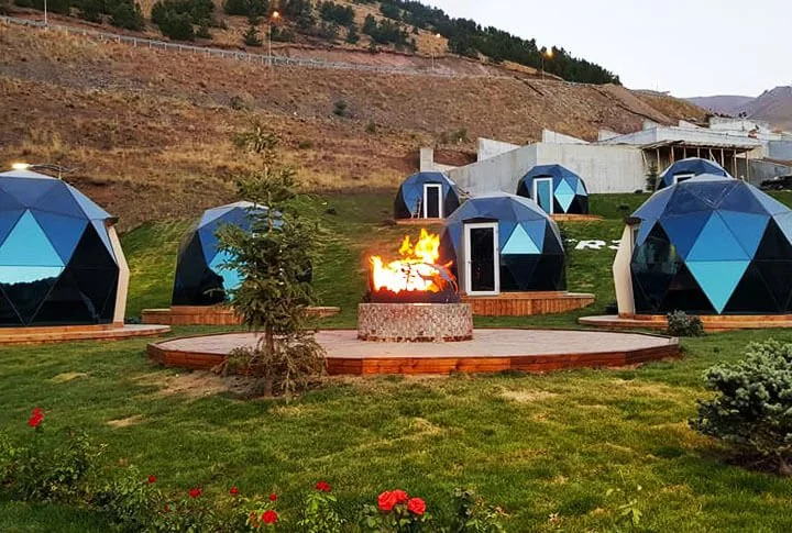 Multiple geodesic dome-shaped tiny homes arranged around an outdoor fire pit on a grassy hillside.