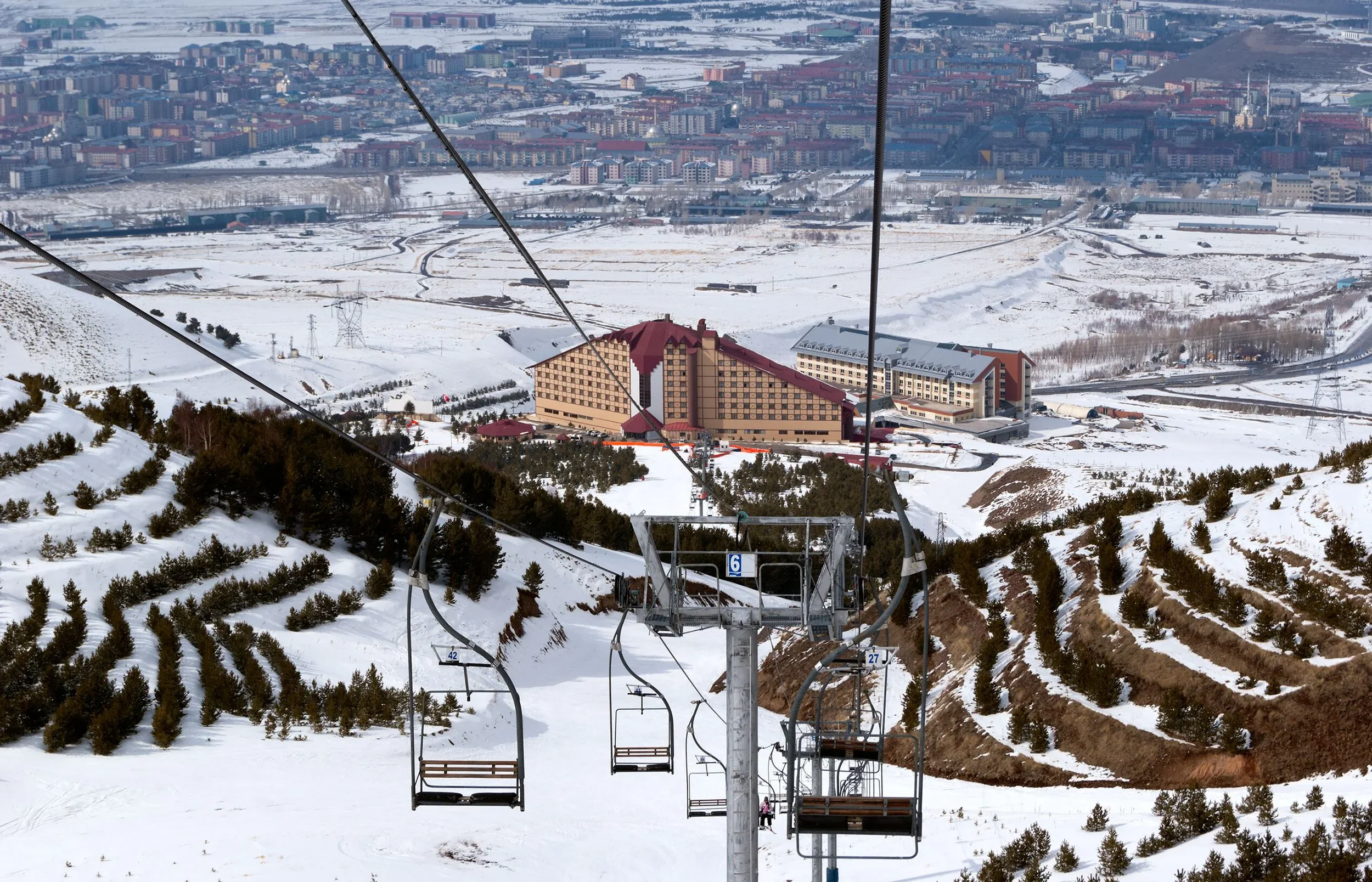 Empty ski lift chairs on a snowy mountain slope with a resort and town in the background.