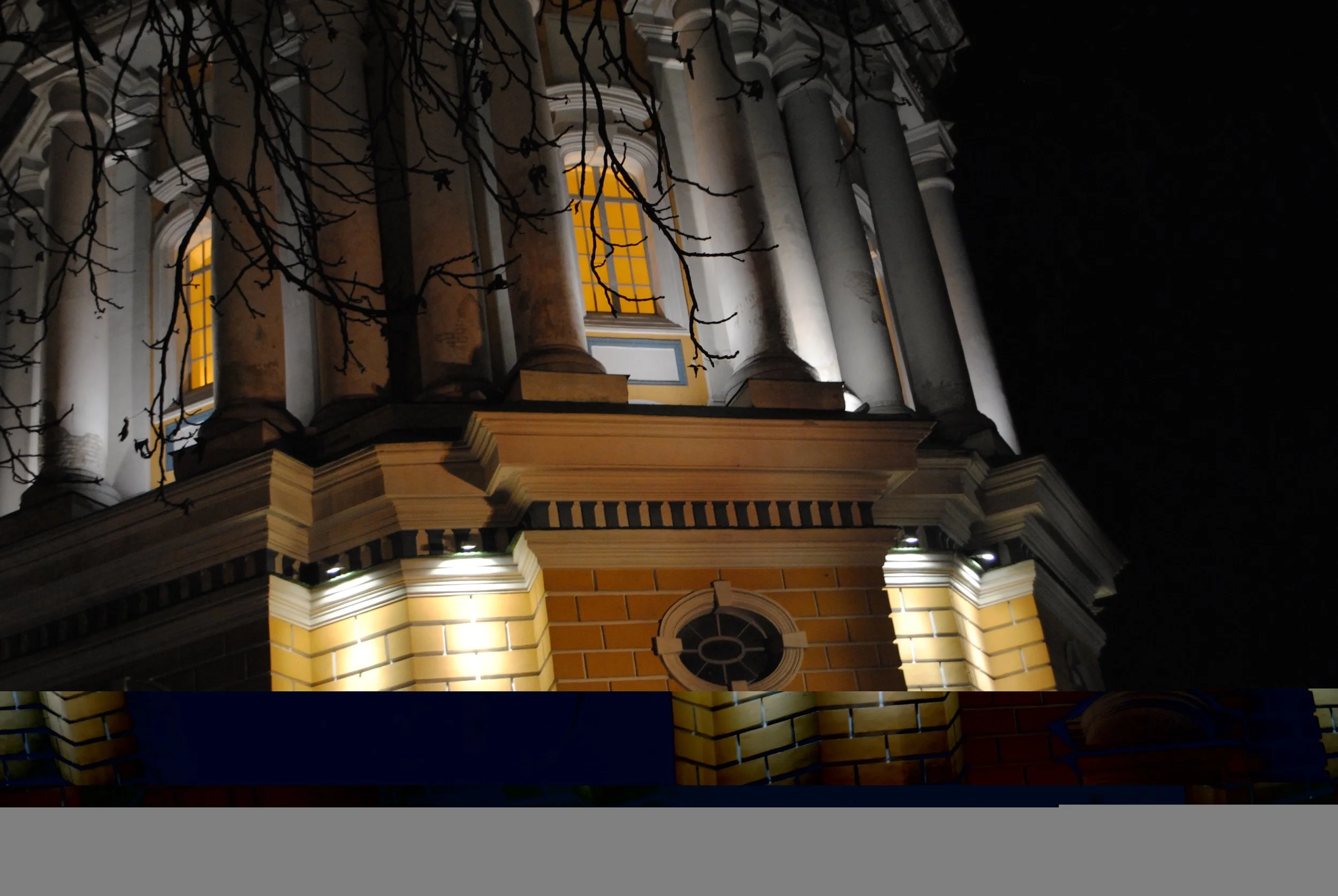 Night view of a church or historic building with illuminated yellow windows, stone columns, brickwork, and a decorative circular window, seen from below with tree branches in the foreground.