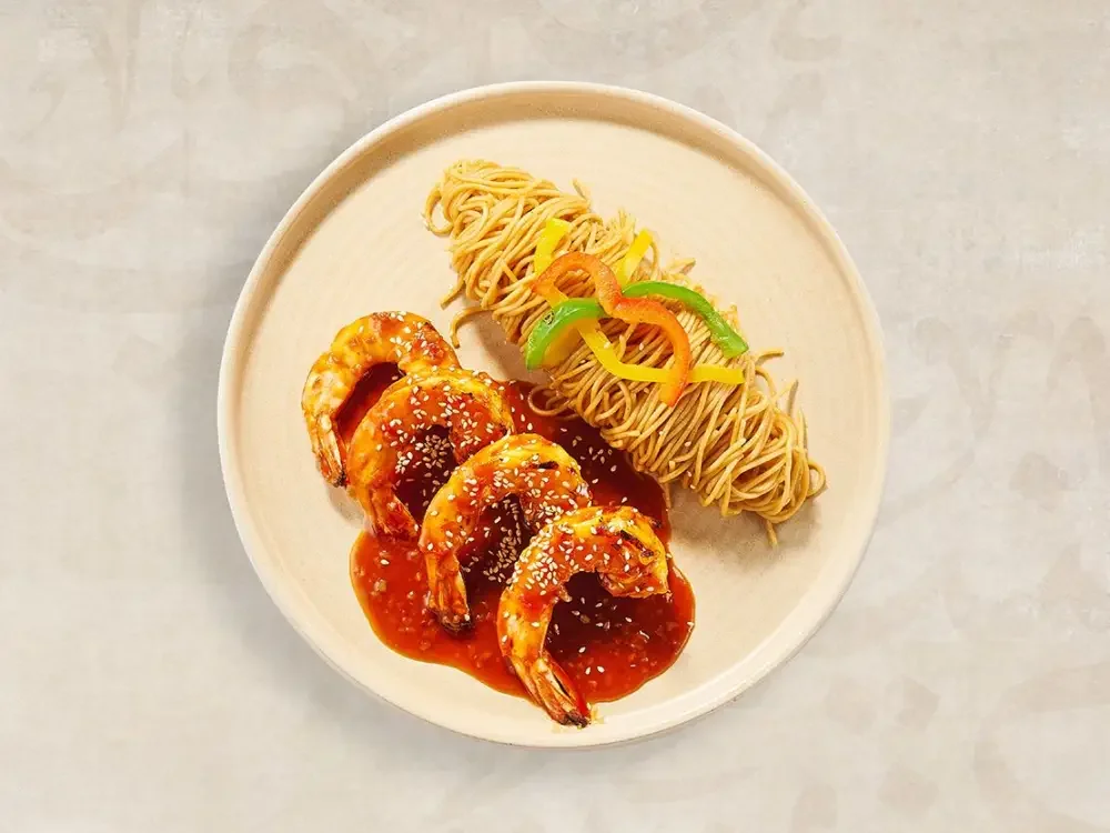 Plate with three grilled shrimp over tomato sauce, sesame seeds, and two types of Asian noodles, garnished with sliced green and yellow bell peppers.