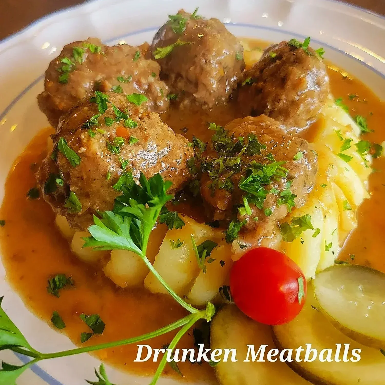A plate of cooked ground meat meatballs in gravy served over mashed potatoes, garnished with chopped parsley and a sprig of parsley, with a cherry tomato and sliced potatoes on the side.