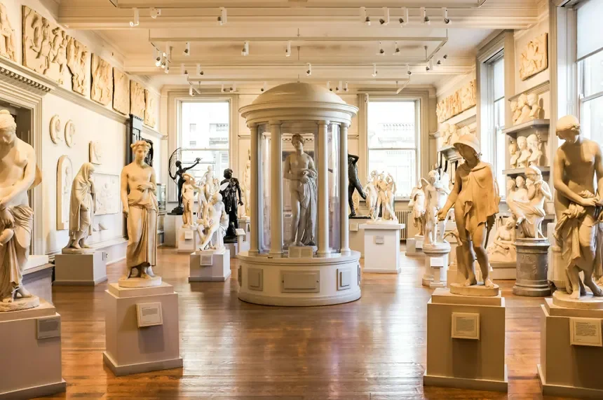 A gallery room filled with white classical sculpture statues, some on pedestals, arranged around a central circular structure. The room has large windows, wooden floors, and artwork displayed on the walls.