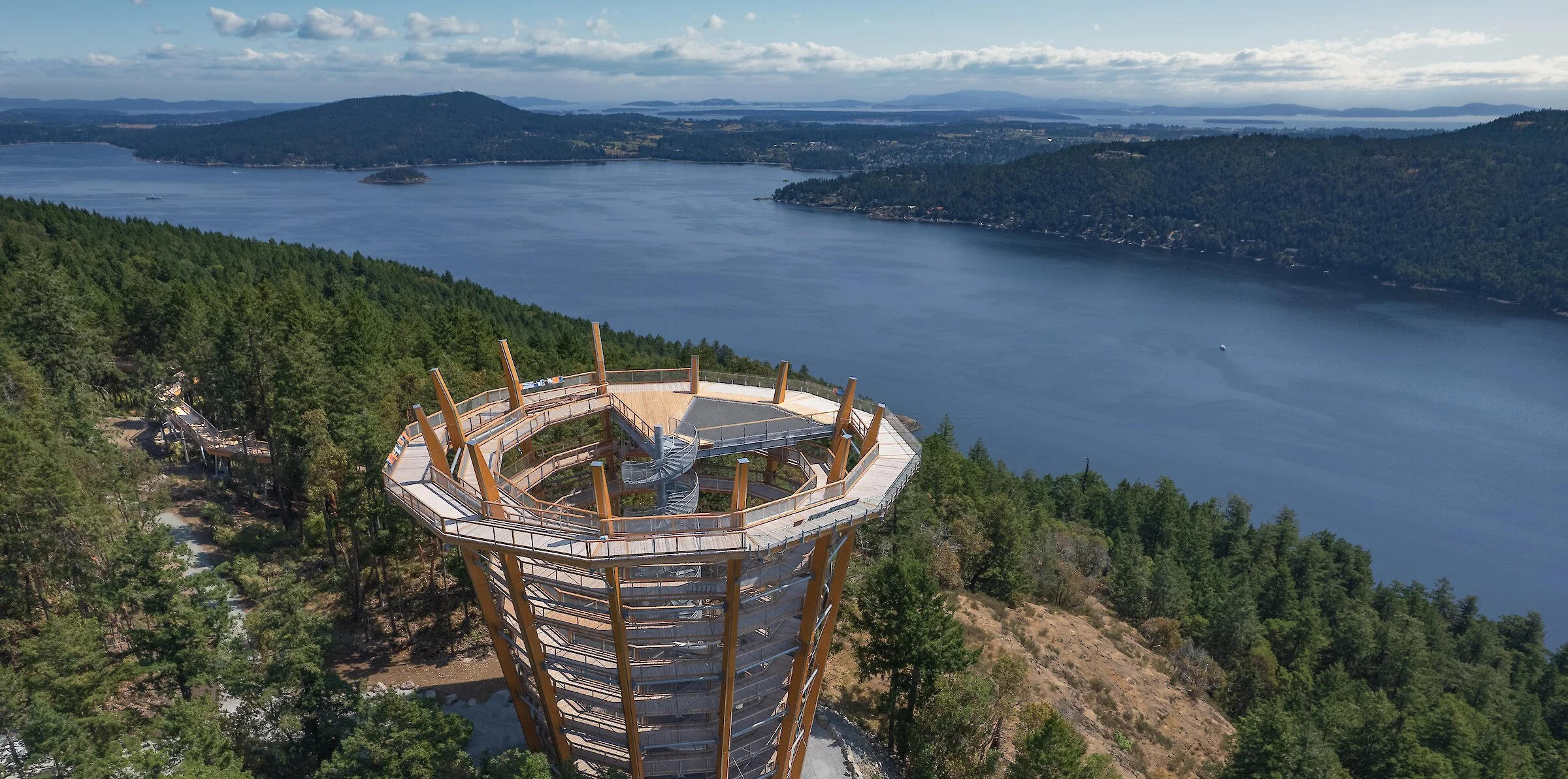 A spiral observation tower on a hillside overlooking a large body of water, surrounded by dense forest and distant mountains, under a partly cloudy sky.