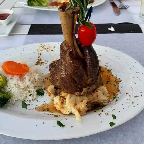 A plate with a cooked lamb shank topped with a cherry and green onion, served with mashed potatoes, rice with vegetables, and broccoli.