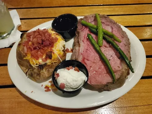 A plate of a baked potato topped with cheese and bacon bits, a large cut of cooked beef with green beans on top, and two small containers of sour cream and dark dipping sauce, served on a wooden table.