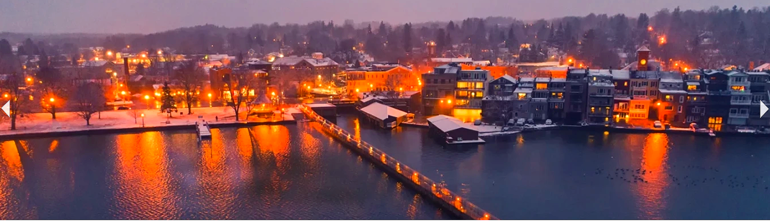 A snowy townscape at dusk with buildings, trees, and a waterway reflecting orange city lights.