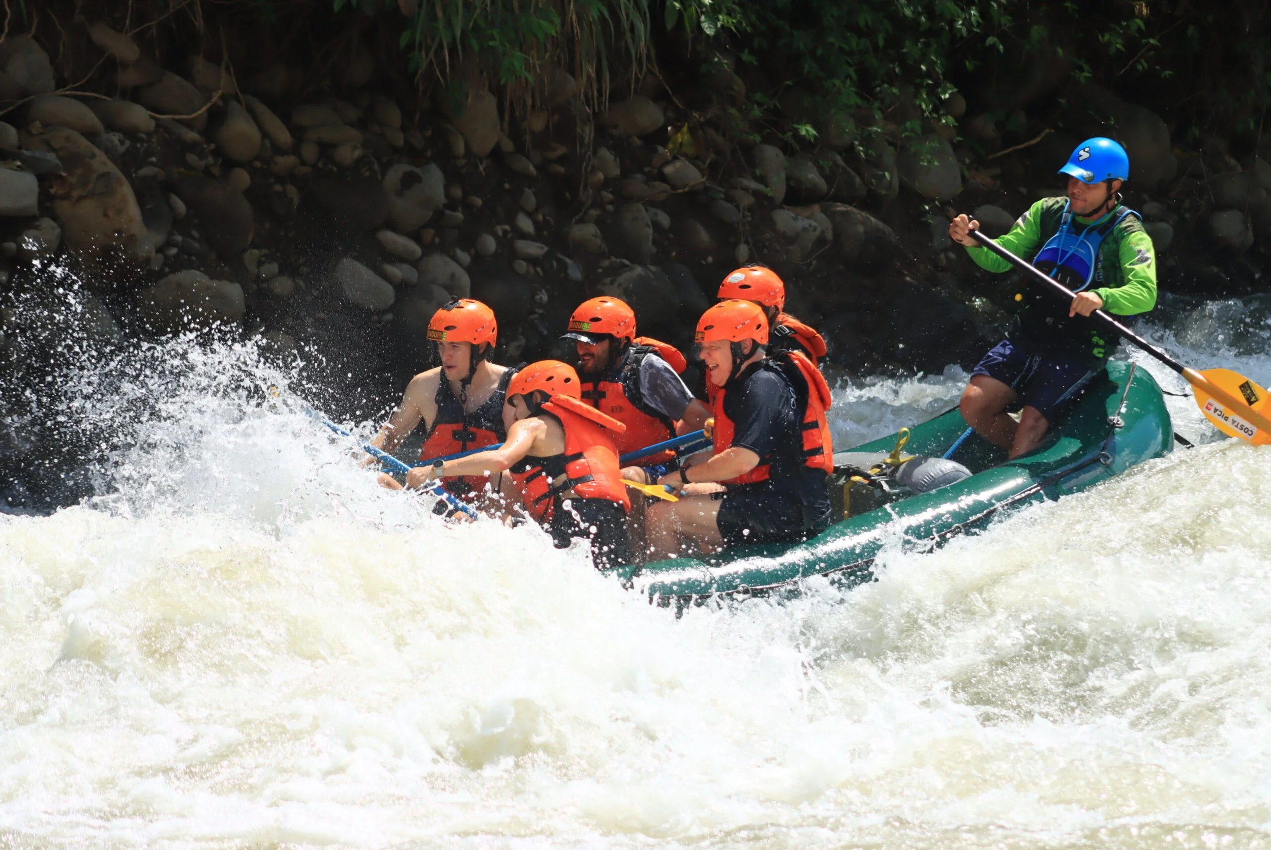 Group of five people white water rafting on a river, wearing orange helmets and life jackets, with an instructor wearing a blue helmet and green jacket paddling at the back.