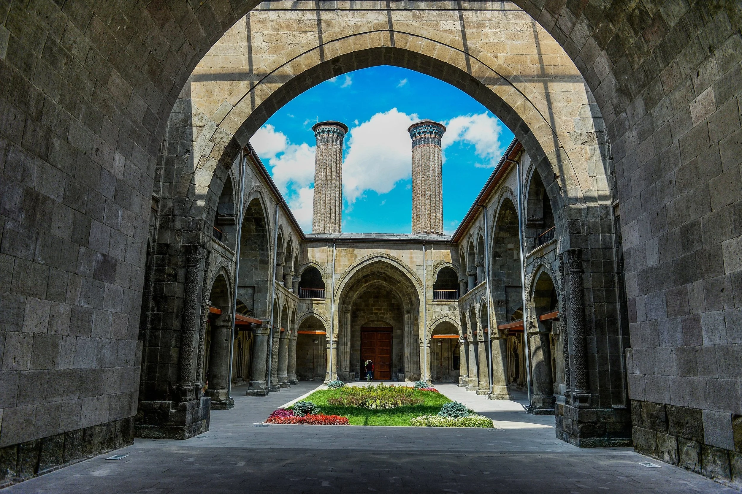 A view through an arched stone gateway into a courtyard with a garden, stone building walls, and two tall stone chimneys or towers against a partly cloudy blue sky.