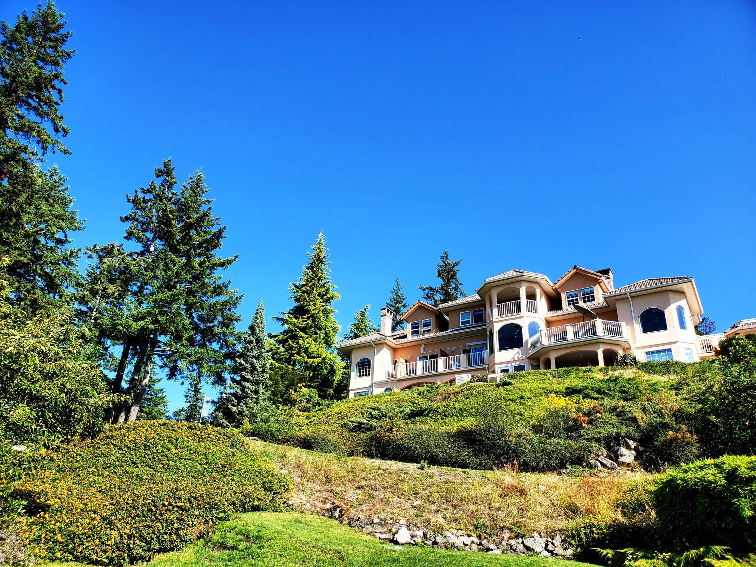Large beige house on a hill surrounded by green trees and bushes under a clear blue sky.