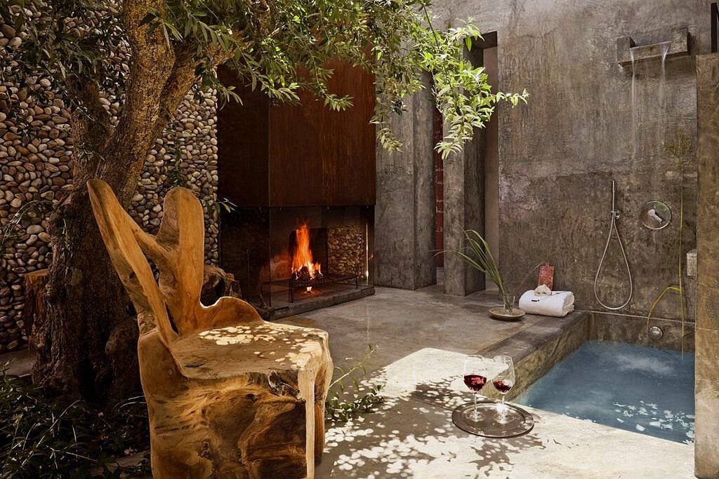 An outdoor spa area with a wooden chair, a small pool, a fire in a fireplace, a tray with two glasses of red wine, and various plants and towels.