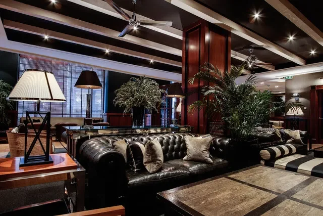 Luxurious hotel lobby with black leather sofas, decorative pillows, large potted plants, table lamps, and modern ceiling lighting.
