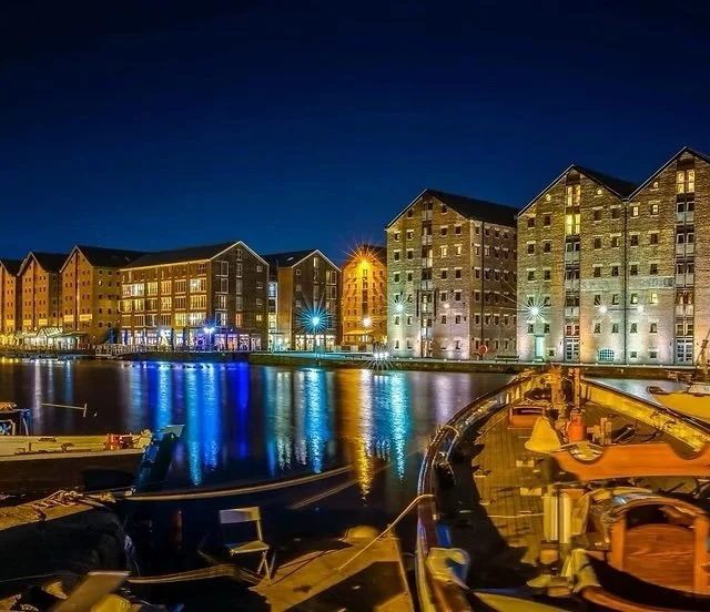 Night view of a waterfront with illuminated multi-story brick buildings and boats docked in the foreground.