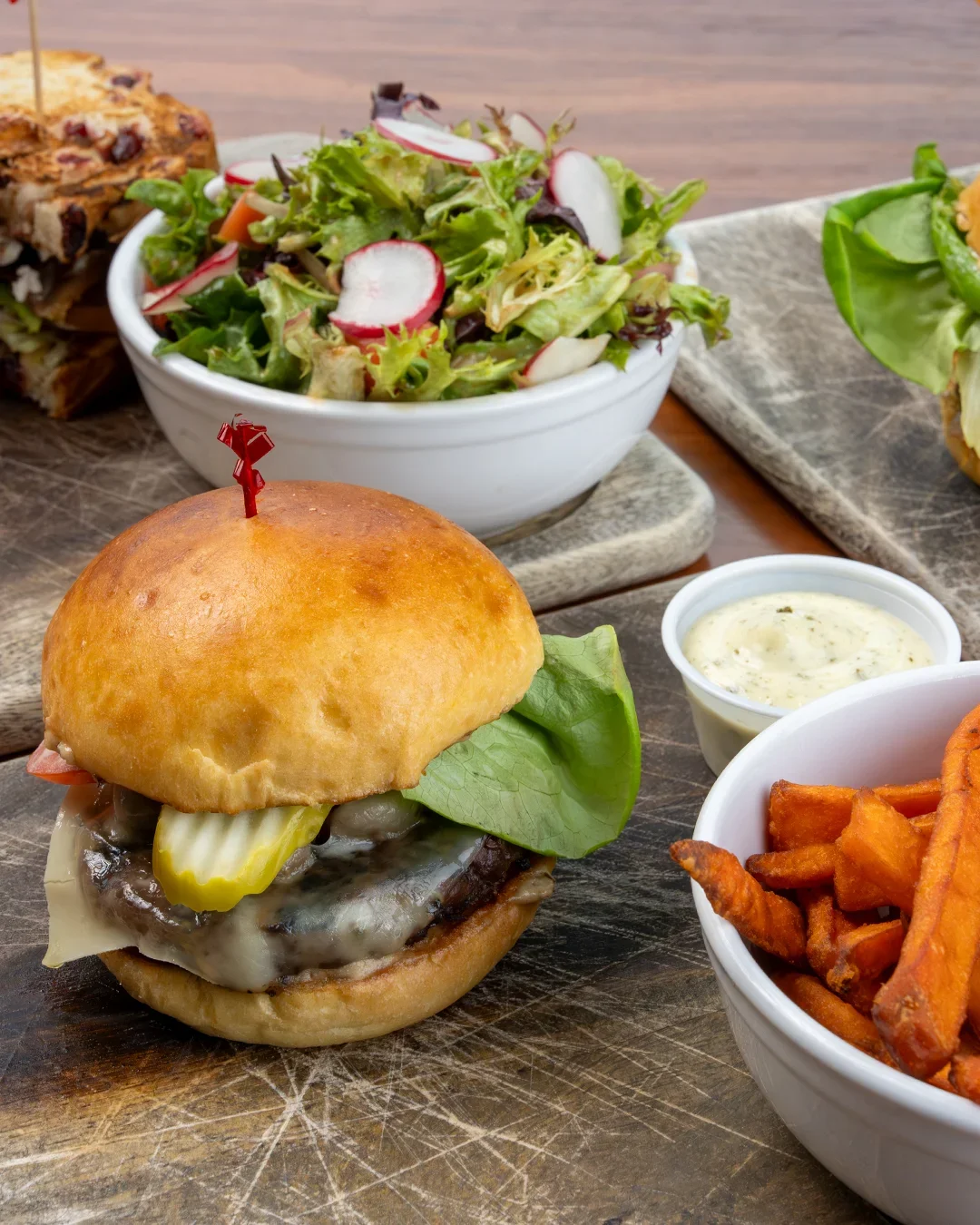 A cheeseburger with pickles, lettuce, and a sandwich bun, accompanied by a side of sweet potato fries with dipping sauce, and a bowl of mixed greens salad with radish slices.