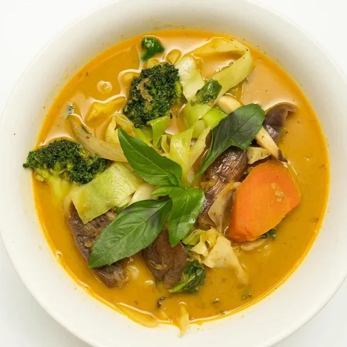 A bowl of vegetable curry with broccoli, carrot, cabbage, eggplant, and fresh basil leaves in a yellow, flavorful broth.