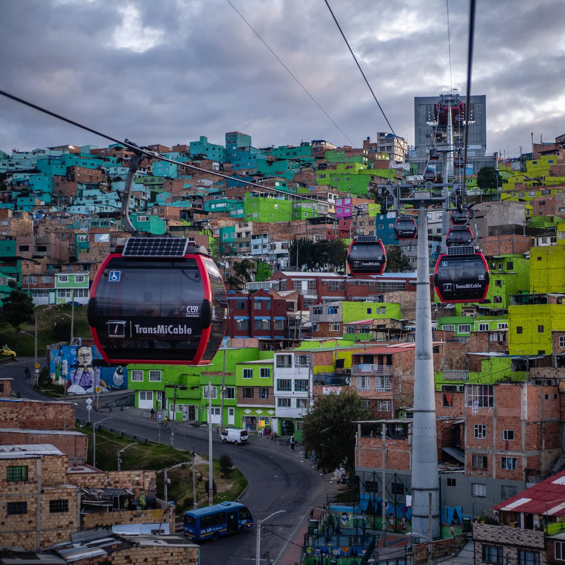 A hillside view of a neighborhood with colorful houses, a cable car system in the foreground, and a cloudy sky overhead.