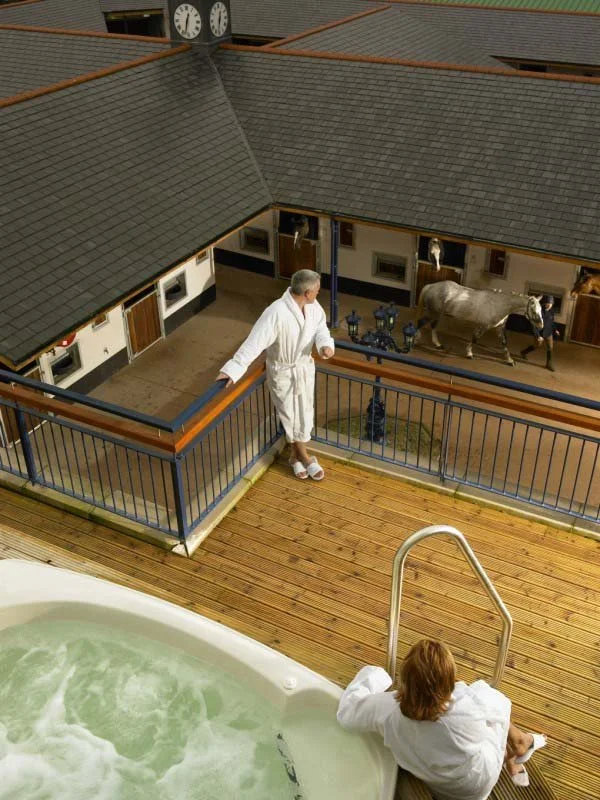 A man in a white robe standing on a balcony overlooking a stable with a horse and a woman in a white robe sitting by a hot tub.