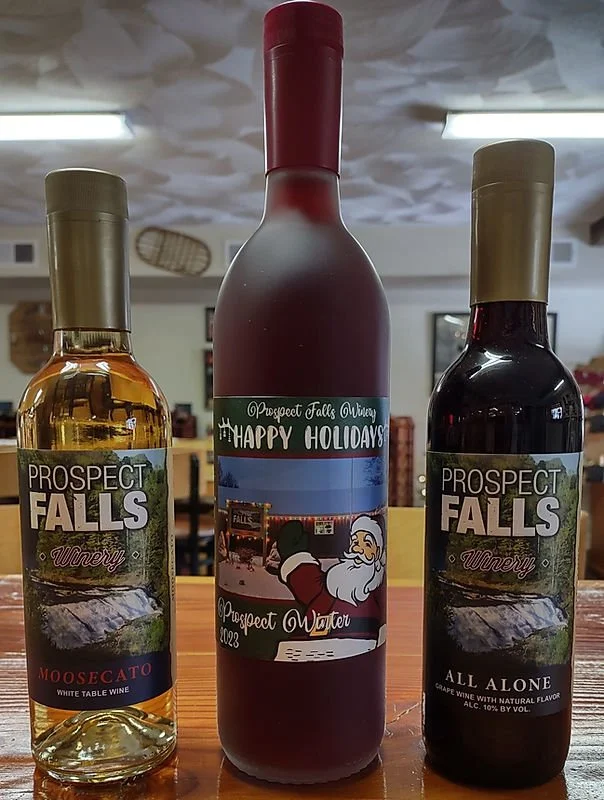 Three bottles of wine from Prospect Falls Winery on a wooden table. The left bottle is a Moscato, the middle bottle is a holiday-themed water label, and the right bottle is a Grape wine labeled All Alone.