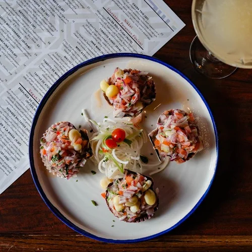 Plate of five small food appetizers topped with chopped meat, vegetables, and cheese, garnished with a cherry tomato and onion slices, next to a glass of white wine and restaurant menus on a wooden table.