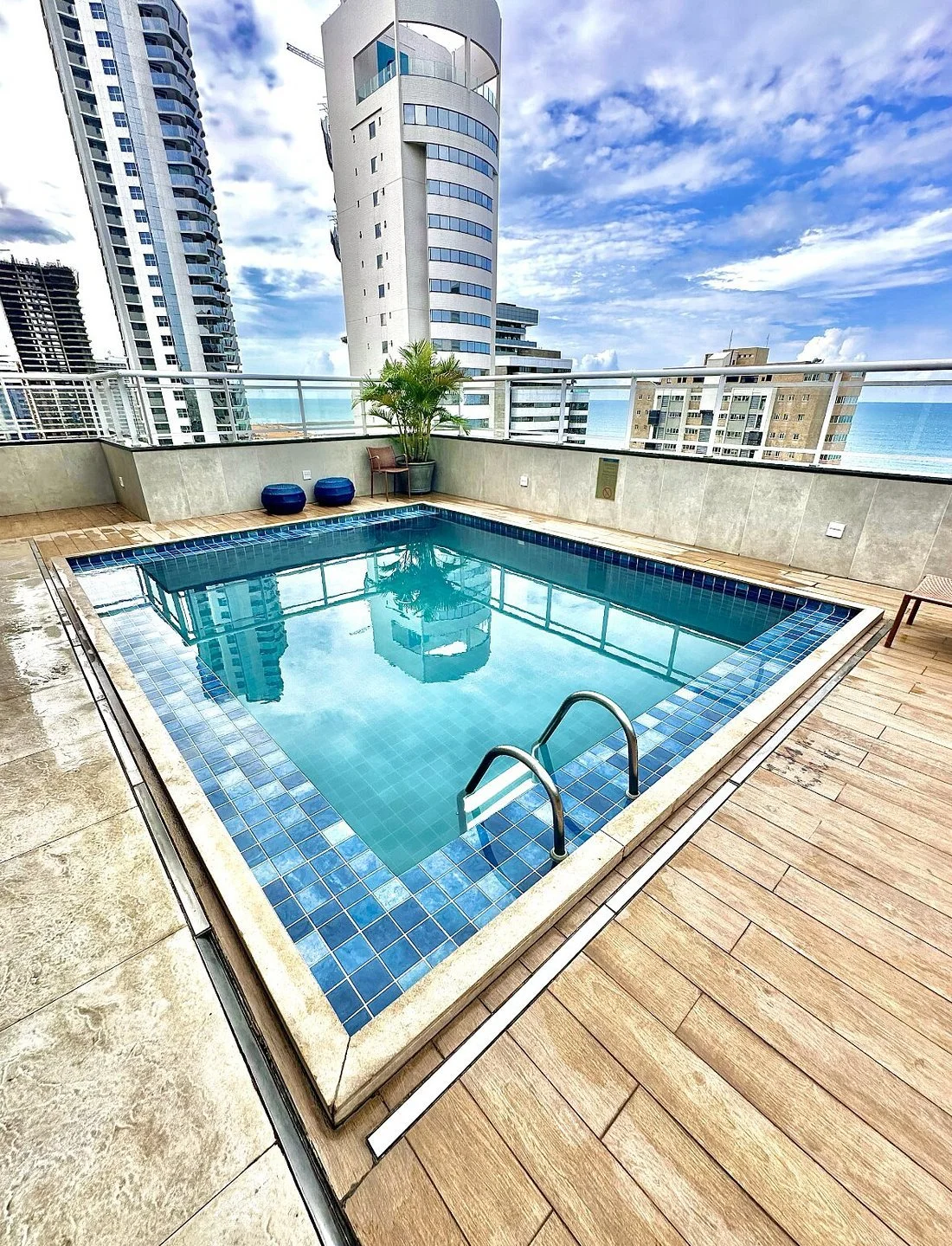 Rooftop swimming pool with city skyline and ocean view, with potted plants, two blue stools, a chair, and a wooden deck