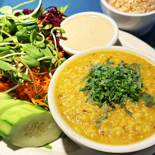 A plate of Indian food featuring a bowl of yellow lentil soup garnished with cilantro, along with fresh cucumber slices, mixed greens, shredded carrots, and beetroot salad, accompanied by small bowls of sauces or chutneys.
