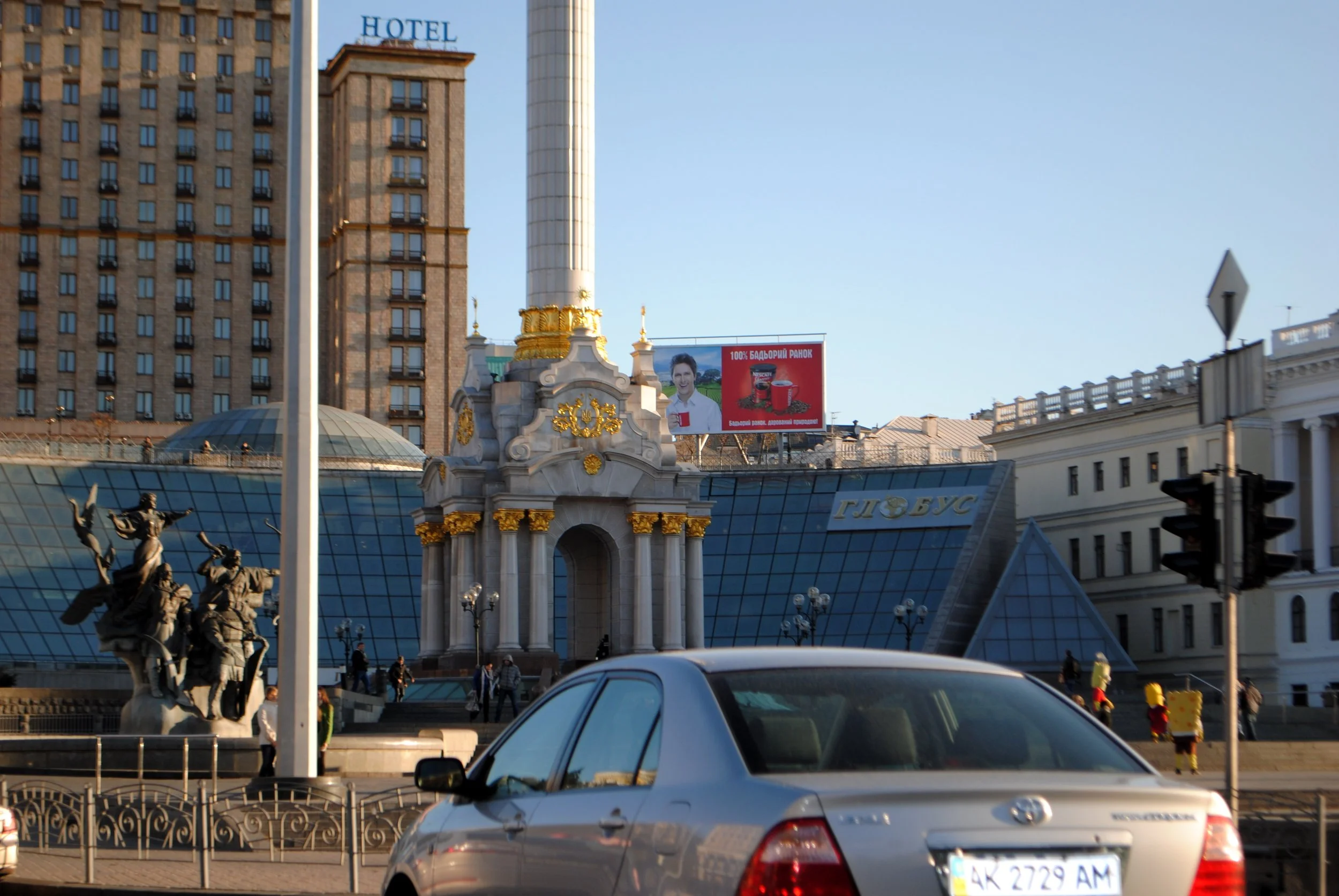 A cityscape with a historic building topped with a white column and gold decorations, modern high-rise buildings in the background, a billboard with a smiling person and red packaging, and sculptures of soldiers in front of the building. Cars are vis