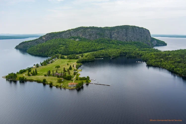 Aerial view of a large lake with an island featuring green grass, trees, and a few buildings, with a steep cliff and forested hillside in the background.
