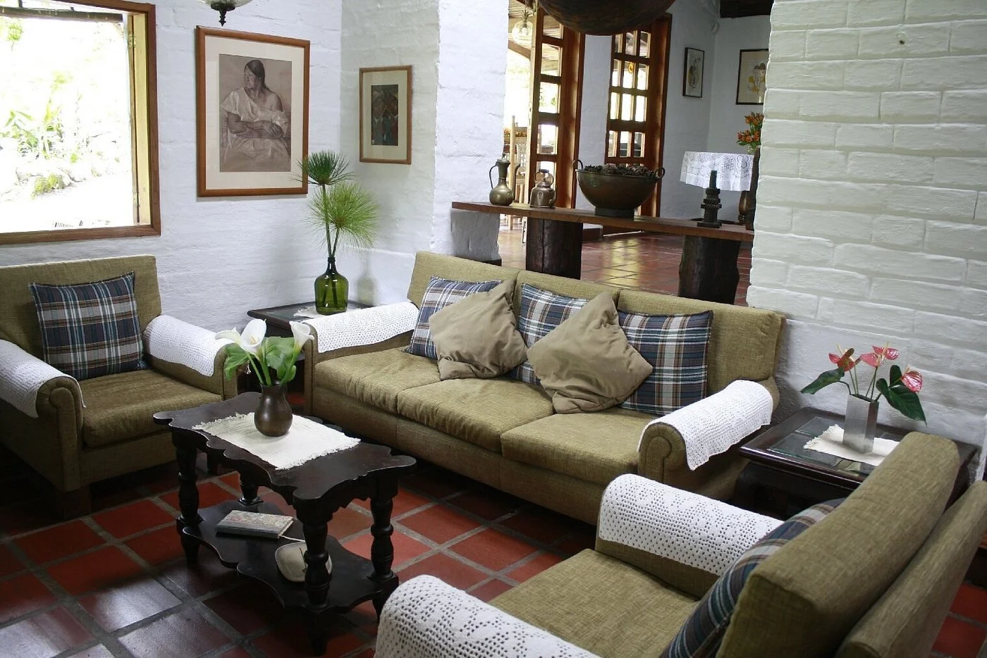 Living room with green sofas, plaid pillows, wooden coffee table with flowers, white brick walls, artwork, and decorative items.
