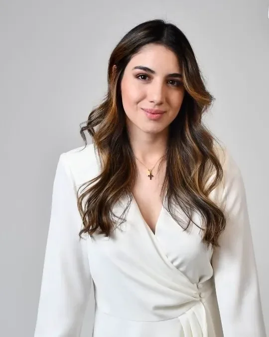 Portrait of a young woman with long wavy brown hair, wearing a white wrap dress and a gold necklace, smiling softly against a light gray background.