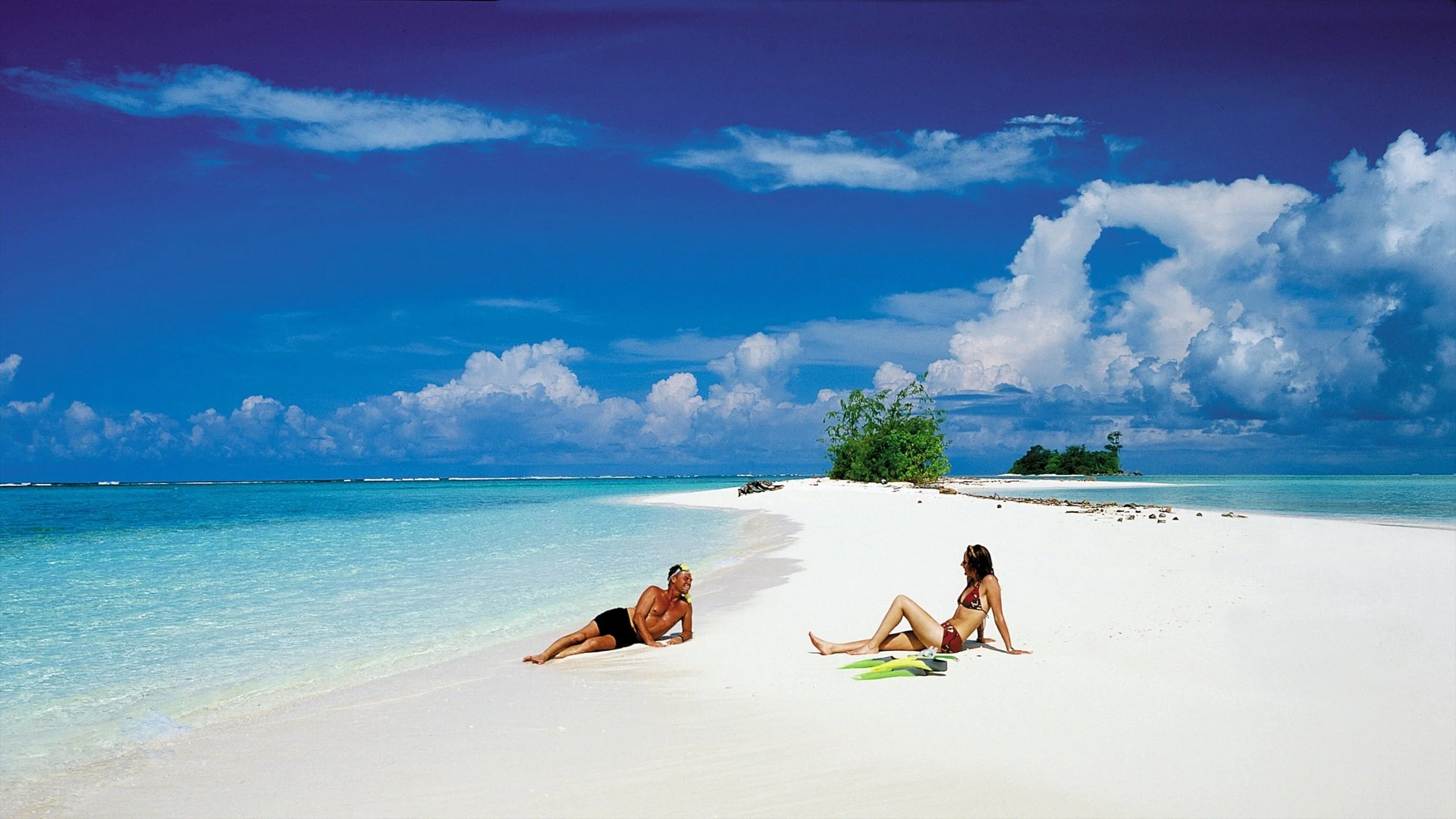A man and woman relaxing on a white sandy beach with turquoise water and green island in the background under a partly cloudy sky. Kiev November 2011, Marina Parrish ©