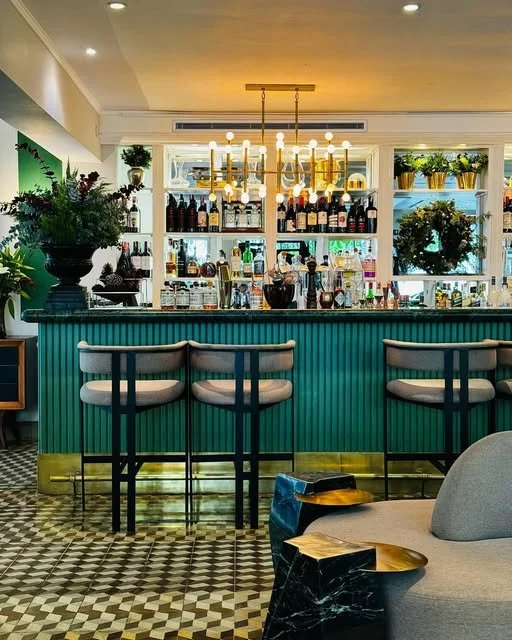 A stylish bar with a teal counter and three white barstools, decorated with plants, bottles, and a chandelier.