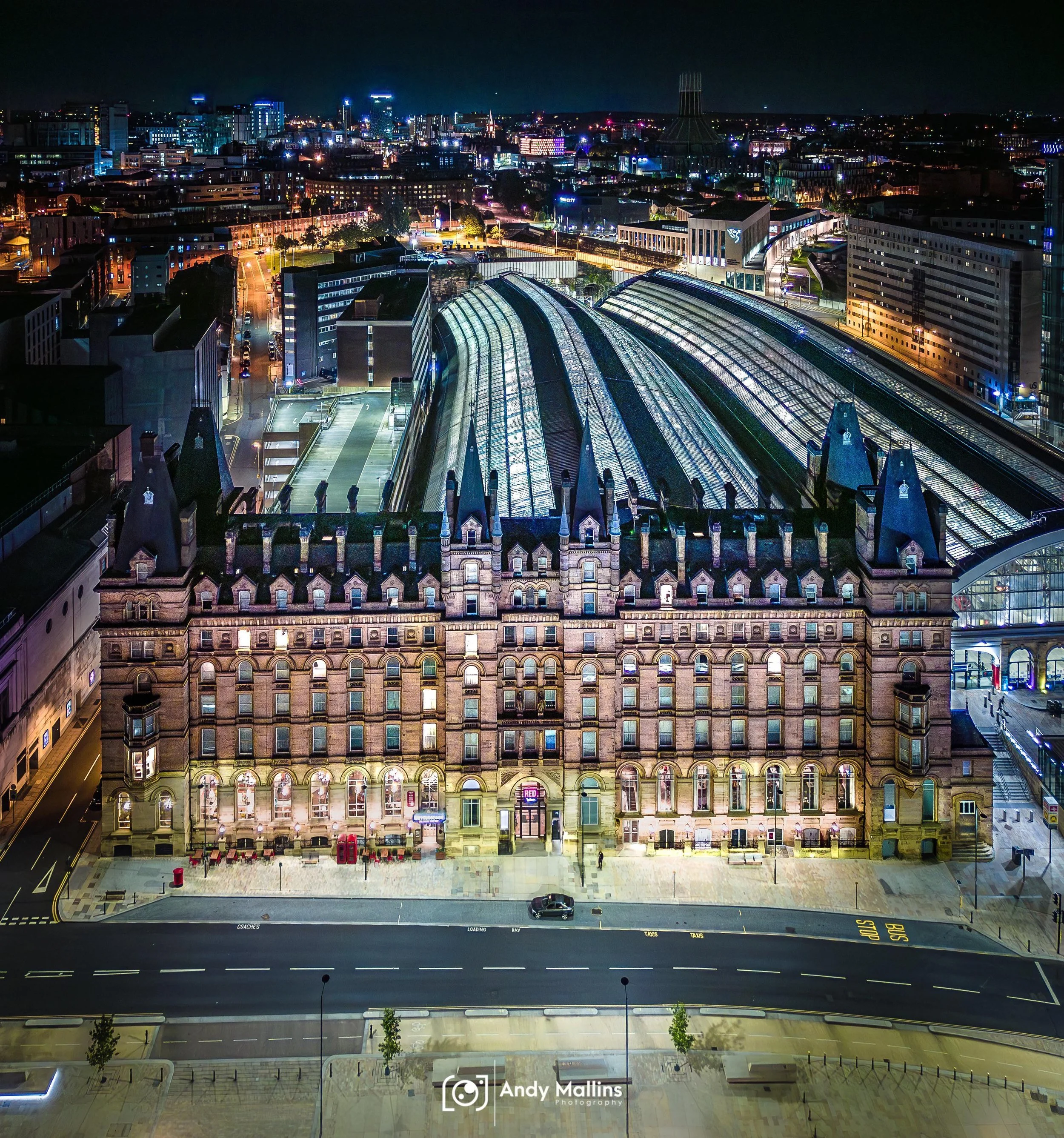 Nighttime cityscape with illuminated buildings, streets, and a large train station with glass roof in the foreground. City skyline with tall buildings in the background.