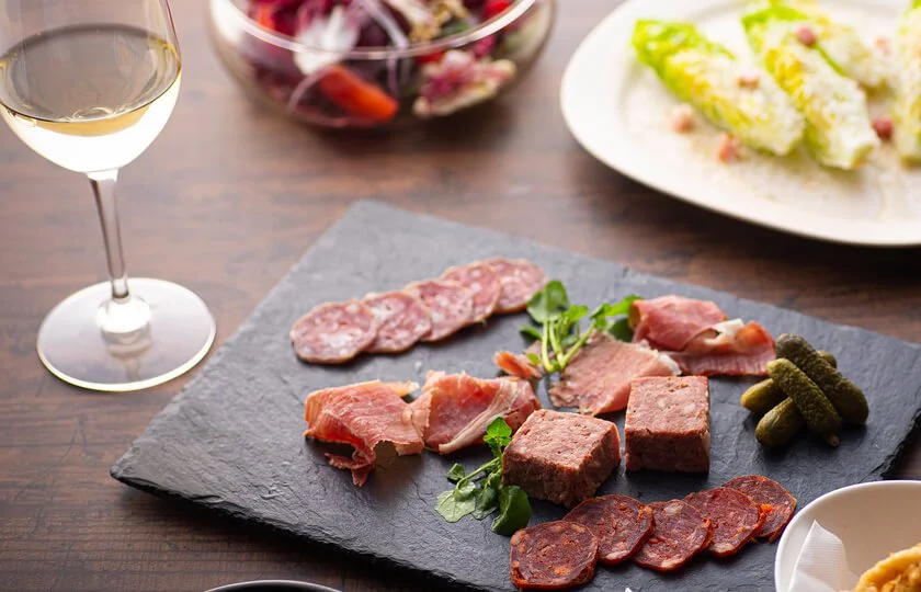 Charcuterie board with meats, pickles, and garnishes, a glass of white wine, and side dishes in the background on a wooden table.