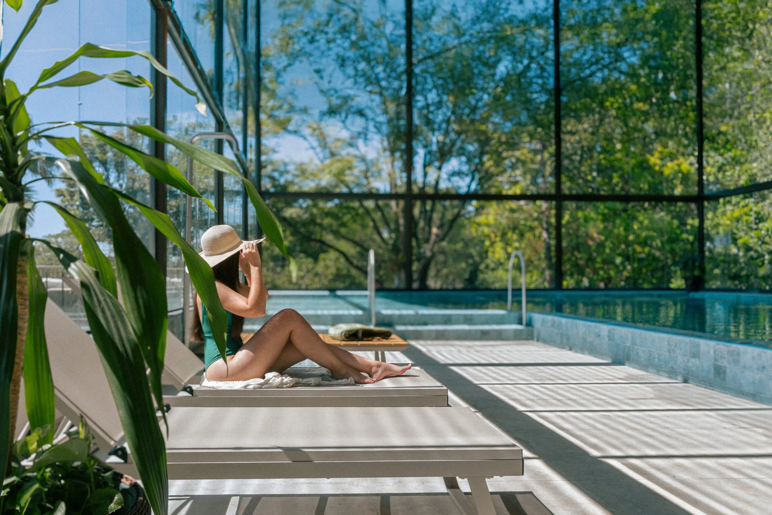 A woman sitting by an outdoor swimming pool, wearing a sun hat and swimsuit, shaded by green plants, with a glass building and trees in the background.