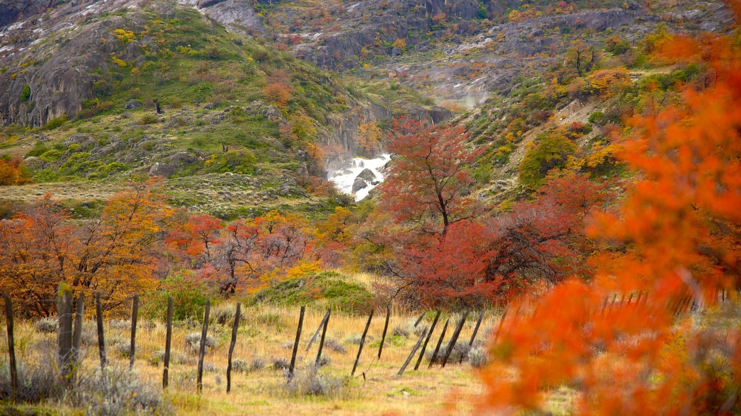 A scenic landscape of a mountainous area during autumn, featuring trees with vibrant red, orange, yellow, and green leaves, a rushing waterfall in the middle, and a fence in the foreground.