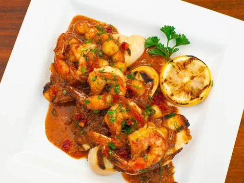 Seafood dish featuring grilled fish topped with shrimp in tomato sauce, garnished with chopped chives, a lemon half with grill marks, and a sprig of parsley on a white square plate.