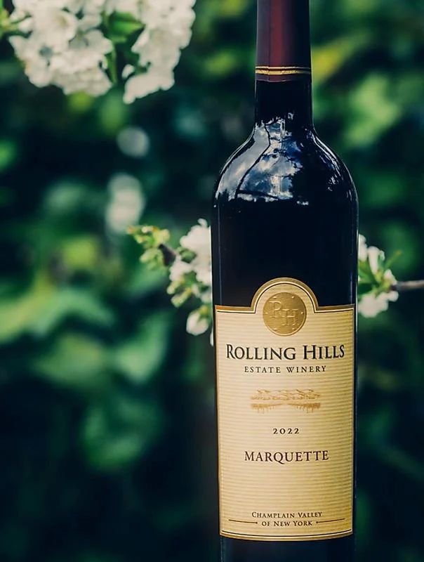 A bottle of Rolling Hills estate wine with a beige label, gold emblem, and black and gold cap, set outdoors with green foliage in the background.