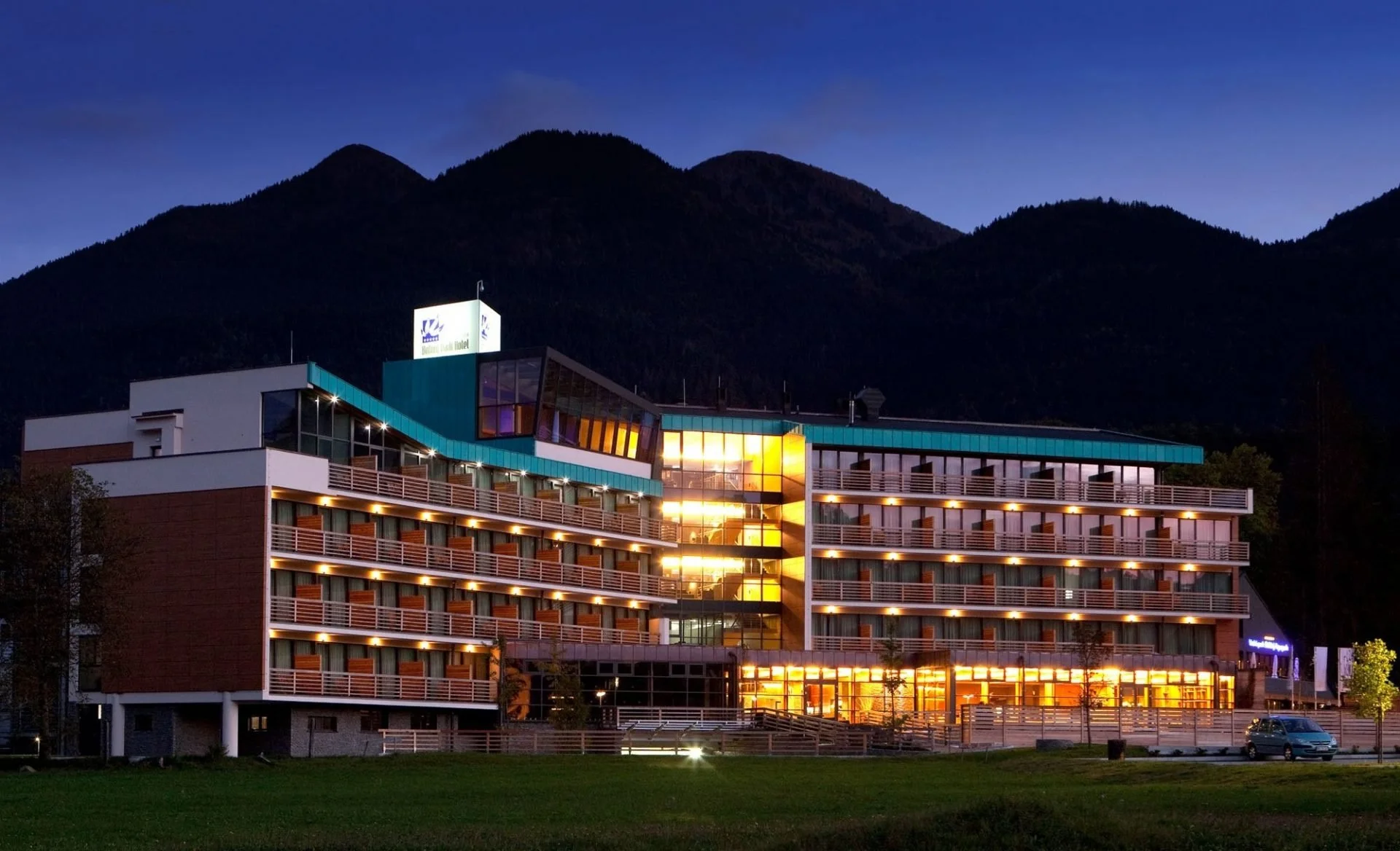 Modern hotel building with multiple balconies illuminated at evening, set against a mountainous background.