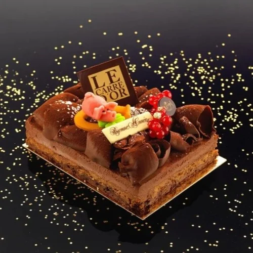 Chocolate cake decorated with assorted candies, a chocolate plaque, and chocolate curls, placed on a white tray with a black background decorated with gold sprinkles.