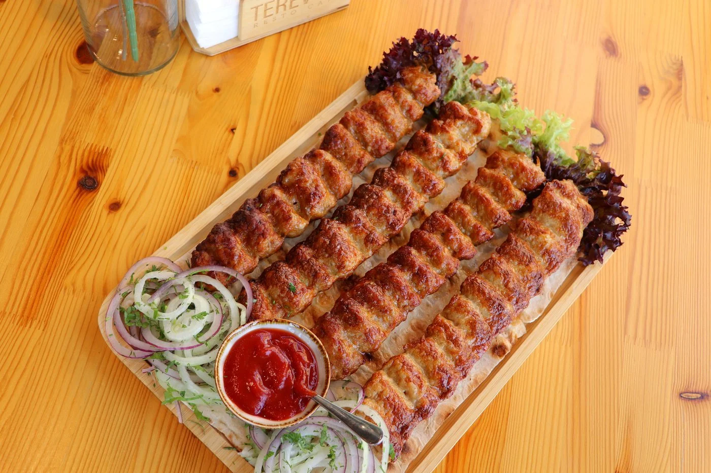 Four grilled skewers of meat on a wooden platter, garnished with lettuce, sliced onions, and a small bowl of ketchup, on a wooden table.