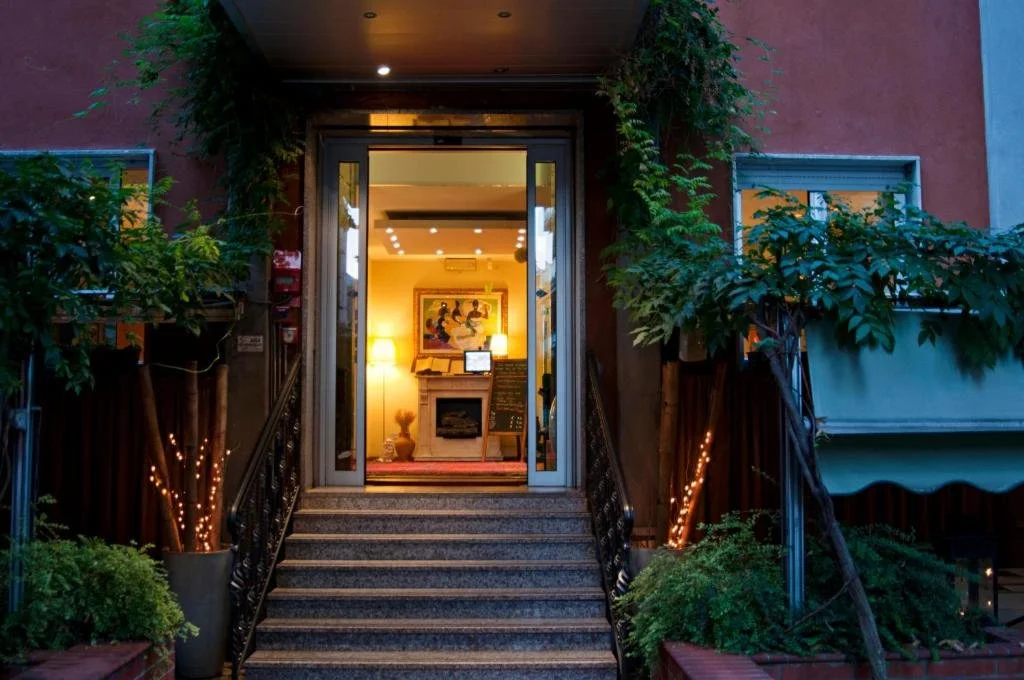 The entrance to a cozy house or cafe is lit with warm yellow lights, with stairs leading up to a glass door. Green plants and small string lights decorate the sides of the stairs.