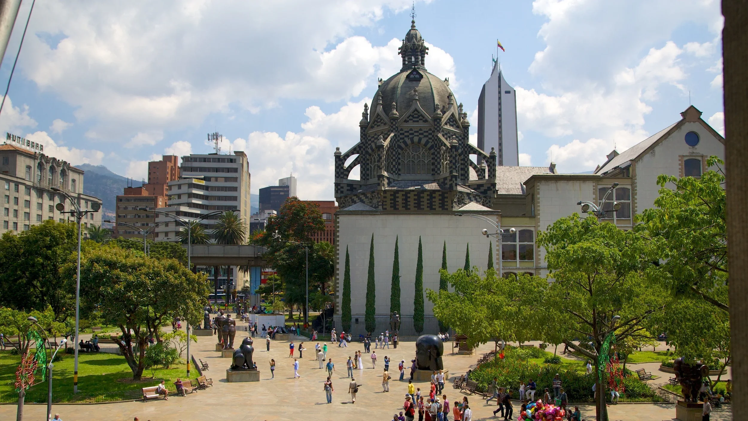 Cityscape with a historic church in the foreground, surrounded by modern buildings and green trees, with people walking and sitting in a park.