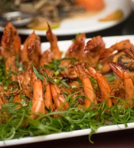 Close-up of cooked shrimp garnished with green herbs on a white platter with fresh green garnishes in the foreground.