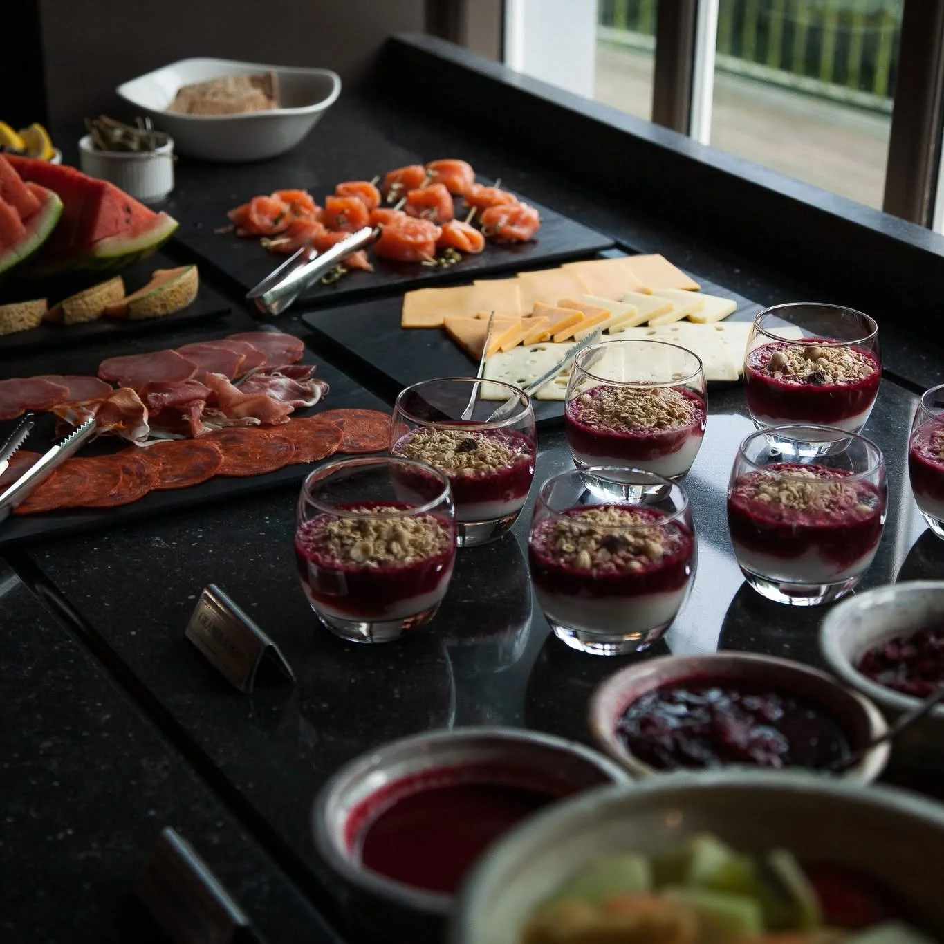 Assorted breakfast food items on a black countertop, including yogurt parfaits topped with granola and berries, sliced watermelon, cheese, sliced charcuterie, and croissants, with a window in the background.