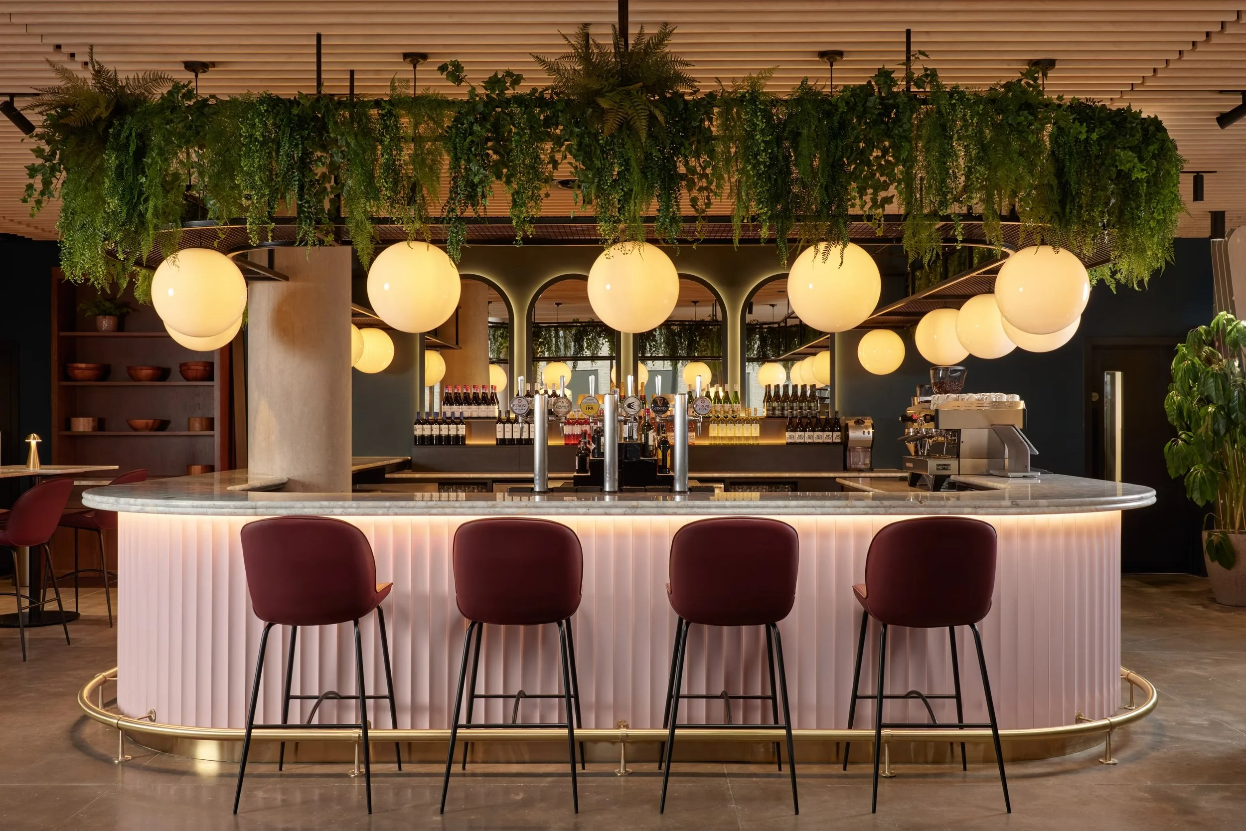 Modern bar with hanging globe lights, lush green plants above the bar, and chairs around a curved bar counter.